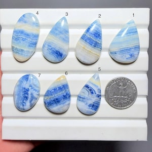 May include: Seven polished blue and white banded gemstones arranged on a white display tray, numbered one through seven. The stones feature varied oval and teardrop shapes with distinct horizontal layers. A US quarter coin is included for scale.