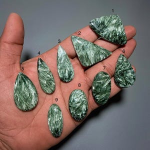 Puede incluir: Colección de nueve piedras preciosas de serafinita verde en varias formas, incluyendo triángulos, gotas y óvalos. Las piedras se muestran en una mano sobre un fondo gris, mostrando sus patrones y texturas únicos.