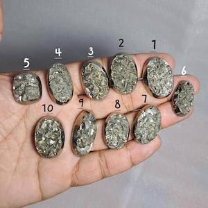 Pode incluir: Uma coleção de dez pedras preciosas de pirita em várias formas, incluindo ovais e quadradas, com uma cor dourada metálica. As pedras são engastadas em engastes de metal prateado e são exibidas em uma mão.