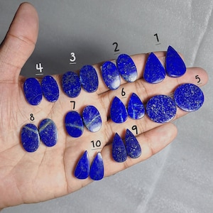 May include: A collection of ten pairs of polished lapis lazuli gemstones in various shapes, including oval, teardrop, and round. The deep blue stones have gold flecks, and are displayed on a light background.
