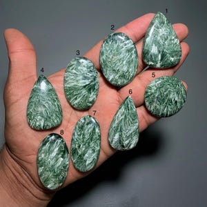 May include: A collection of eight green seraphinite gemstones in various shapes, including oval, teardrop, and round. The stones display a feathery, textured pattern. The gemstones are displayed on a hand against a gray background.
