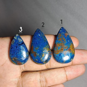Puede incluir: Tres piedras preciosas pulidas en forma de lágrima. Las piedras son principalmente de un azul profundo con vetas marrones y doradas. Las piedras preciosas están numeradas 1, 2 y 3. Las piedras se muestran sobre un fondo neutro.
