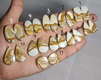 May include: A collection of eleven polished jasper gemstones in various shapes, including ovals, teardrops, and rectangles. The stones feature a mix of tan, brown, and white colors with unique patterns. The gemstones are displayed on a light background.
