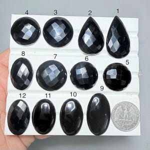 Può includere: Assortimento di dodici pietre preziose nere di varie forme, tra cui goccia, ovale e rotonda, esposte su una scheda bianca. Alcune pietre hanno superfici sfaccettate, altre lisce. Una moneta da un quarto di dollaro USA è inclusa per riferimento dimensionale.