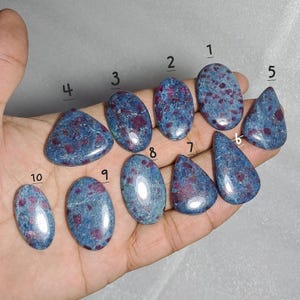 Puede incluir: Una colección de diez piedras preciosas pulidas, cada una con una forma única. Las piedras presentan un patrón moteado de tonos rojo oscuro y azul. Las piedras preciosas se sostienen en una mano, mostrando sus formas y colores individuales.