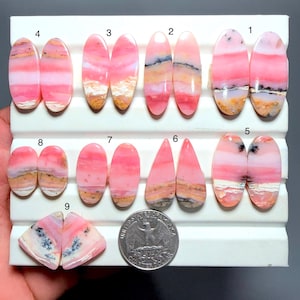 Puede incluir: Una colección de cabujones de ópalo rosa pulidos dispuestos en pares numerados sobre un expositor blanco, con patrones de bandas en tonos rosa, blanco y bronceado, algunas piedras muestran dendritas negras, junto a una moneda para escala.