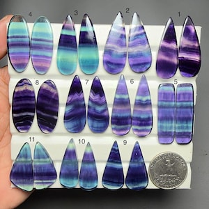 May include: A collection of polished fluorite cabochons in various shapes, including ovals, teardrops, and rectangles, featuring banded purple, blue, and green patterns. Each pair is numbered from 1 to 11, with a US quarter coin for scale.