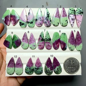 May include: A collection of polished ruby zoisite cabochons in various shapes, including teardrop and oval, arranged in rows on a white display tray. Each stone features a mix of green zoisite and magenta ruby, with a US quarter coin for scale.