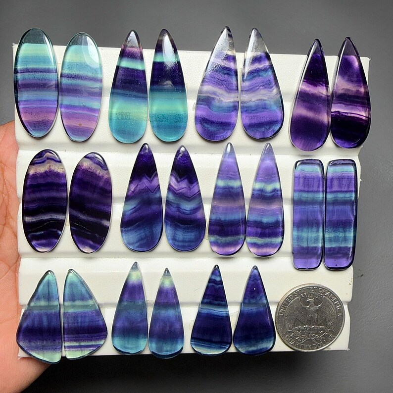 May include: A collection of polished fluorite cabochons in various shapes including ovals, teardrops, and rectangles, featuring banded patterns of purple, blue, and green. A United States quarter coin is included for scale on the white display board.