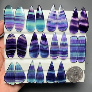 May include: A collection of polished fluorite cabochons in various shapes including ovals, teardrops, and rectangles, featuring banded patterns of purple, blue, and teal. A United States quarter coin is included for scale on the white display board.