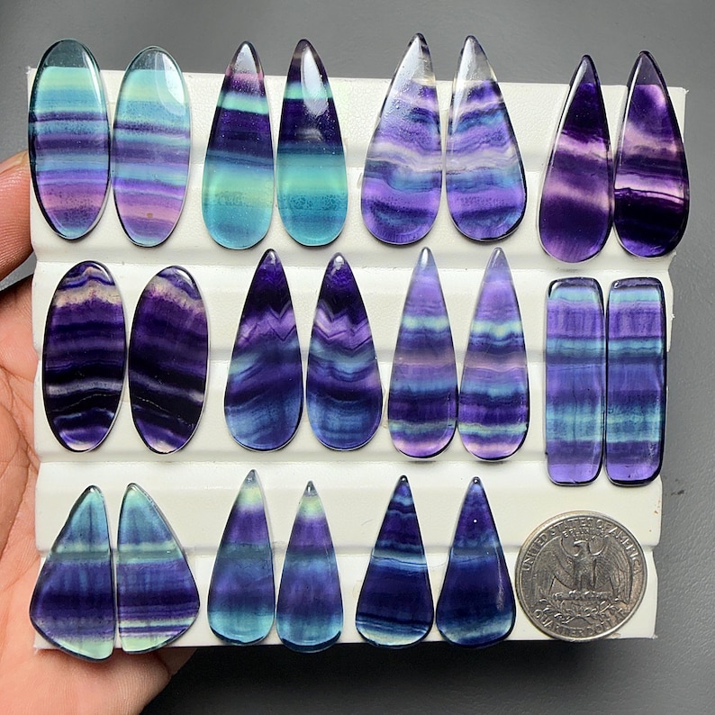 May include: A collection of polished fluorite cabochons in various shapes including ovals, teardrops, and rectangles, featuring banded patterns of purple, blue, and teal. A United States quarter coin is included for scale on the white display board.