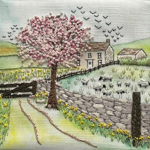 May include: Embroidered landscape depicting a flowering tree, stone wall, and a path leading to a house. Sheep graze in the field. Black birds fly in the sky. The artwork is on a square, off-white fabric.