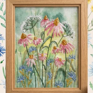 May include: A framed embroidery art piece featuring a botanical scene with pink coneflowers, small blue cornflowers, and white lace-like flowers stitched onto a light green and yellow fabric background, enclosed in a light-toned wooden frame.