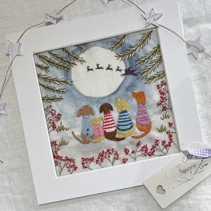 May include: Embroidered picture in a white frame. The image shows five dogs in sweaters, looking at a full moon with reindeer. The scene is set in a snowy landscape with red floral accents. A tag with text and a heart is attached.