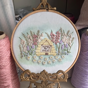 May include: A circular wooden embroidery hoop containing a fabric panel with a hand-stitched design of a yellow beehive, pink and purple wildflowers, and white daisies. A small sign on the hive features the embroidered text Honey For Sale.