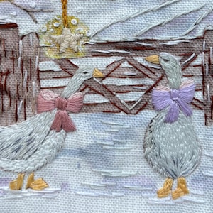 May include: A hand-embroidered scene on white fabric featuring two geese with yellow beaks and feet. One goose wears a pink bow, the other a lavender bow. The background shows a brown wooden fence, a yellow star ornament, and white snow.