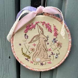 May include: A beige embroidery hoop with a pink ribbon and a brown bunny embroidered in the centre. The bunny is surrounded by pink flowers and green leaves.