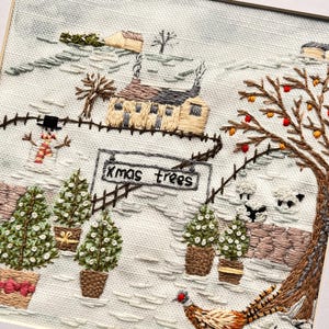 May include: A square embroidery piece on light grey fabric depicting a winter landscape with a house, a snowman, potted evergreen trees, and a sign reading "Xmas trees". A pheasant stands in the foreground near a tree with orange fruit.