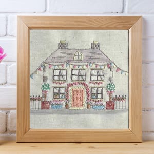 May include: A framed embroidery of a quaint house with a gray roof, adorned with colorful bunting and floral accents. The house features multiple windows, a pink door, and potted plants. A pink tulip in a white vase is on the left.