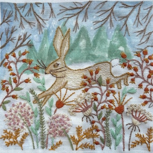 May include: A square fabric panel featuring an embroidered tan rabbit leaping through a winter landscape. The scene includes brown branches, orange berries, and white floral clusters against a backdrop of green mountains and a light blue sky.