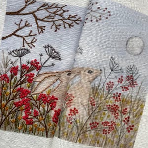 May include: Two embroidered rabbits looking up at a full moon, surrounded by brown, green, and red embroidered foliage.