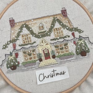 May include: Embroidered Christmas scene showing a detailed cottage with festive decorations and garland. The word "Christmas" is stitched below the cottage. The embroidery is set in a wooden hoop, perfect for holiday decor.