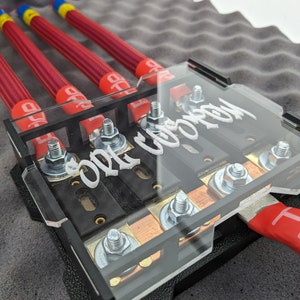 Puede incluir: Un componente eléctrico negro con una cubierta transparente, con el texto "CORE CUSTOM". Tiene múltiples terminales con pernos plateados y cableado rojo y negro. Los cables son rojos con detalles amarillos y azules.