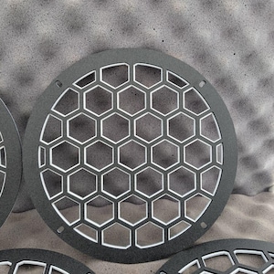 May include: Four dark grey circular speaker grilles with a honeycomb design. The grilles feature a series of hexagonal cutouts and a dark grey finish. Each grille has small mounting holes around the edge for installation.