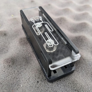 Puede incluir: Un clip de plástico negro y acrílico transparente con un diseño estilizado de figura humana. El clip tiene forma rectangular con una parte superior de acrílico transparente y una base negra. La figura está grabada.