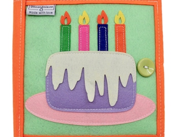 Puede incluir: Un libro de fieltro verde con un borde naranja. El libro presenta una aplicación de fieltro de un pastel morado con glaseado blanco y cuatro velas. Las velas son verdes, rosas, azules y naranjas. El pastel está en un plato rosa. El libro tiene un botón en la parte delantera.