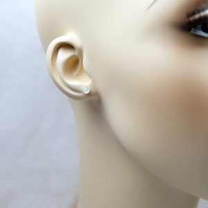 May include: A close-up of a person's ear with a small, white opal stud earring. The earring is set in a gold setting.