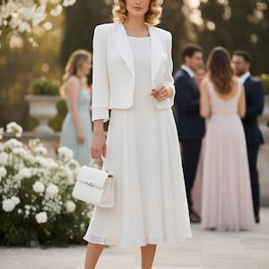 May include: A woman in a white dress, jacket, and hat, holding a white handbag. The dress is knee-length with pleats. The jacket is a cropped style. She is wearing white heels. The background shows a garden setting with other people.