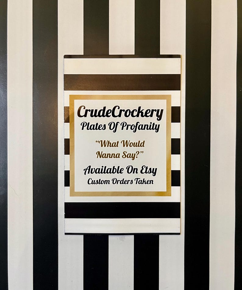 May include: A rectangular sign featuring a black and white horizontal striped pattern with a central gold-bordered white square. The text reads: CrudeCrockery, Plates Of Profanity, &ldquo;What Would Nanna Say?&rdquo;, Available On Etsy, Custom Orders Taken.