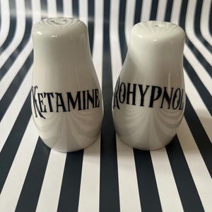 May include: Two white ceramic salt and pepper shakers with a curved shape. The text "KETAMINE" and "ROHYPNOL" are printed in black on the shakers. The background is a black and white striped pattern.