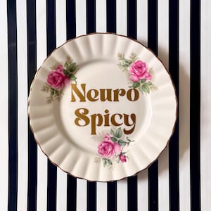 May include: A round, cream-coloured ceramic plate with a scalloped edge and a gold-tone rim. The plate features three clusters of pink roses with green leaves and the text "Neuro Spicy" printed in a bold, gold-coloured serif font in the centre.