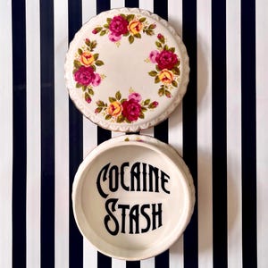 May include: A round ceramic container with a removable lid. The lid features a floral pattern of pink and yellow roses with green leaves on a white background. The interior of the base is white and features the black text "COCAINE STASH" in a bold font.