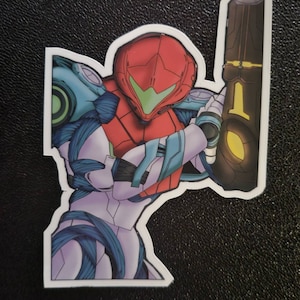 May include: A sticker featuring a cartoon illustration of a video game character, Samus Aran, from the Metroid series. She is wearing a red and blue suit and holding a large weapon. The sticker is white with a black outline.