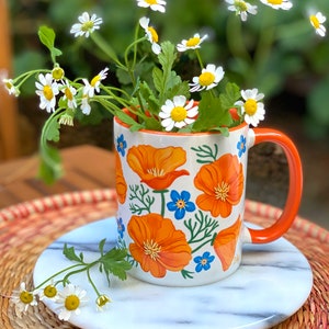 May include: A white ceramic mug with an orange floral design and a bright orange handle. The mug is sitting on a white marble coaster and is surrounded by a bouquet of white daisies.