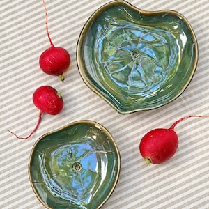 May include: Two ceramic dishes shaped like lily pads with a glossy, variegated green glaze and dark brown edges. The dishes feature a textured, veined surface pattern radiating from a central point. Three bright red radishes are placed nearby.