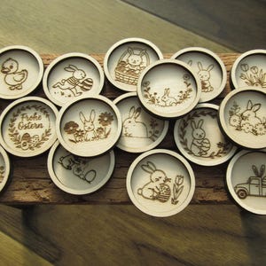 Op de afbeelding: Een verzameling houten paas-onderzetters met lasergeëtste ontwerpen. De onderzetters tonen afbeeldingen van konijnen, eenden, eieren en bloemmotieven. Sommige onderzetters hebben de tekst "Frohe Ostern".