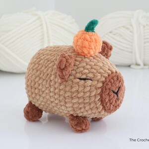 May include: A handmade crochet capybara plush toy in shades of brown, with a small orange pumpkin on its head. The plush has closed eyes and small brown ears and feet. The Crochet Code is written on the bottom right.