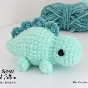 May include: A light teal crocheted dinosaur toy with a darker teal spine and black eyes. The plush toy is made of soft, textured yarn. The image also shows teal yarn in the background. Text on the image reads "No Sew Crochet Pattern".