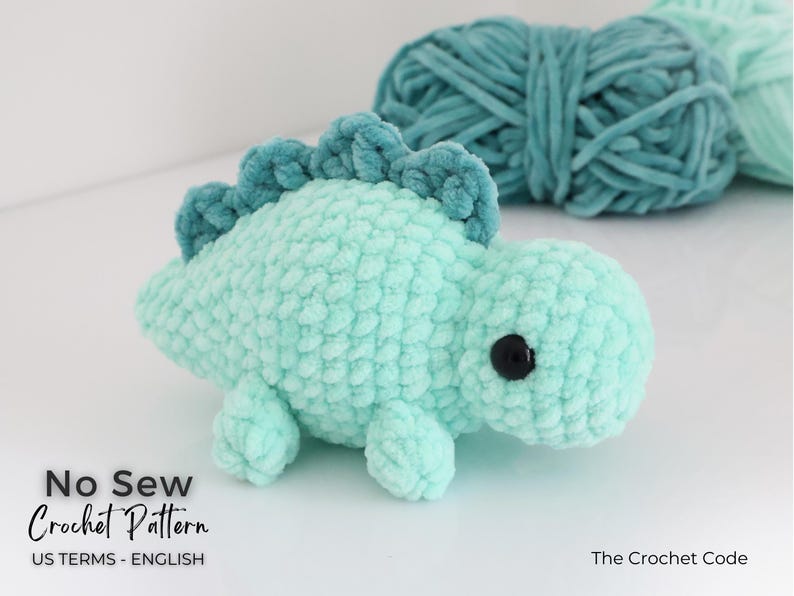 May include: A light teal crocheted dinosaur toy with a darker teal spine and black eyes. The plush toy is made of soft, textured yarn. The image also shows teal yarn in the background. Text on the image reads "No Sew Crochet Pattern".