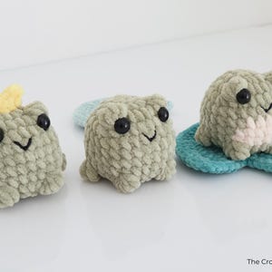 May include: Three handmade crochet frog plushies in shades of green, each with black eyes and a smiling mouth. One frog has a yellow crown. One frog is on a blue lily pad. The Crochet Code is in the bottom right corner.