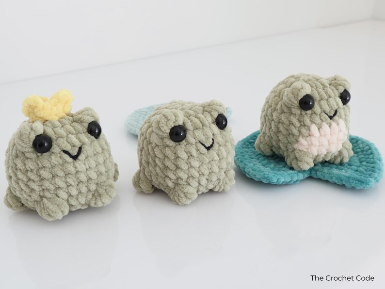 May include: Three handmade crochet frog plushies in shades of green, each with black eyes and a smiling mouth. One frog has a yellow crown. One frog is on a blue lily pad. The Crochet Code is in the bottom right corner.