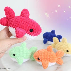 May include: A collection of colorful, crocheted shark plushies. The sharks are in various colors, including pink, blue, yellow, green, and orange, with white bellies and black eyes. The plushies are soft and cuddly, perfect for children.