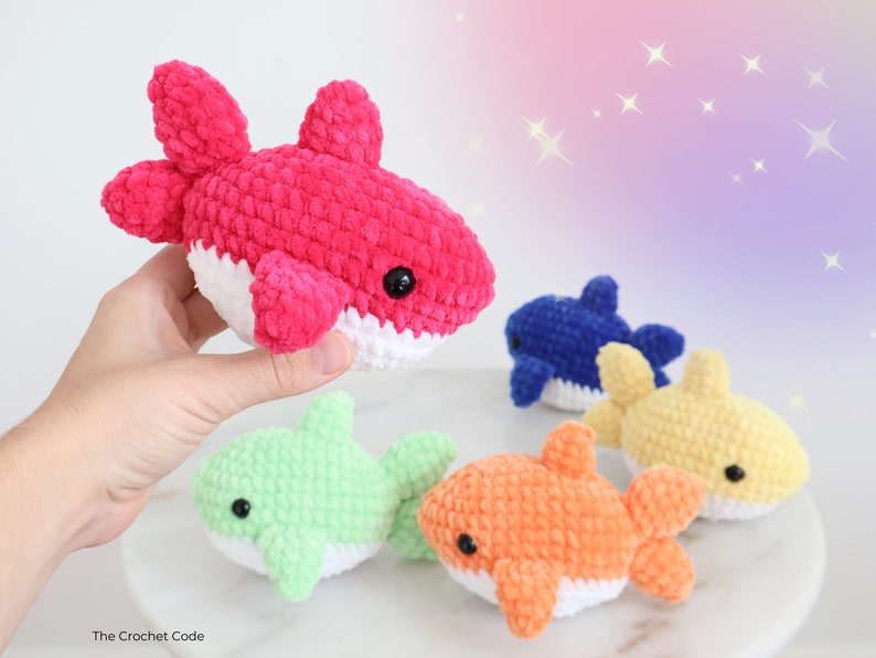 May include: A collection of colorful, crocheted shark plushies. The sharks are in various colors, including pink, blue, yellow, green, and orange, with white bellies and black eyes. The plushies are soft and cuddly, perfect for children.