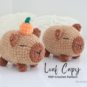 May include: Two crocheted capybara figures in shades of tan and brown, one with a small orange pumpkin on its head. The figures have closed eyes and are labeled "Loaf Capy" with "PDF Crochet Pattern" below. White yarn is in the background.