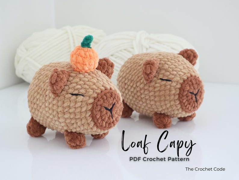 May include: Two crocheted capybara figures in shades of tan and brown, one with a small orange pumpkin on its head. The figures have closed eyes and are labeled "Loaf Capy" with "PDF Crochet Pattern" below. White yarn is in the background.