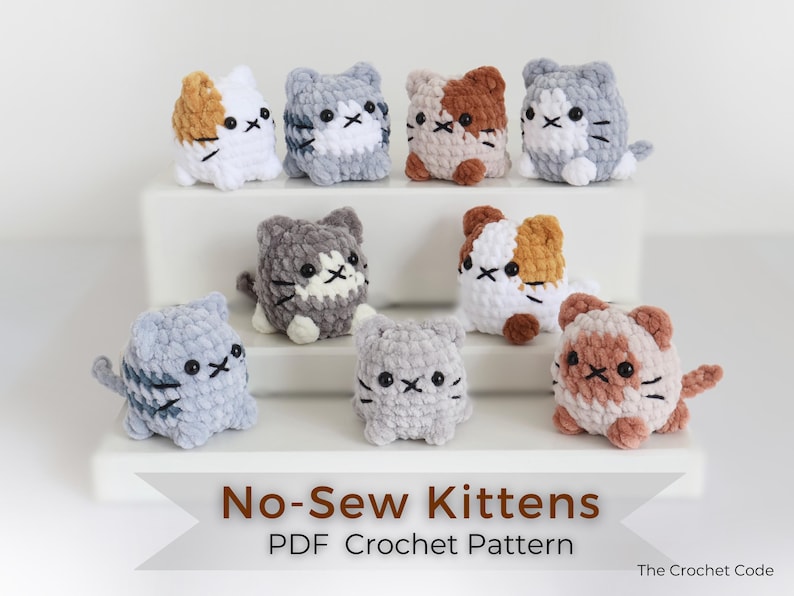 May include: A collection of eight crocheted kitten figurines in various colors, including white, gray, and brown. Each kitten has black embroidered facial features. The image includes text that reads "No-Sew Kittens PDF Crochet Pattern".
